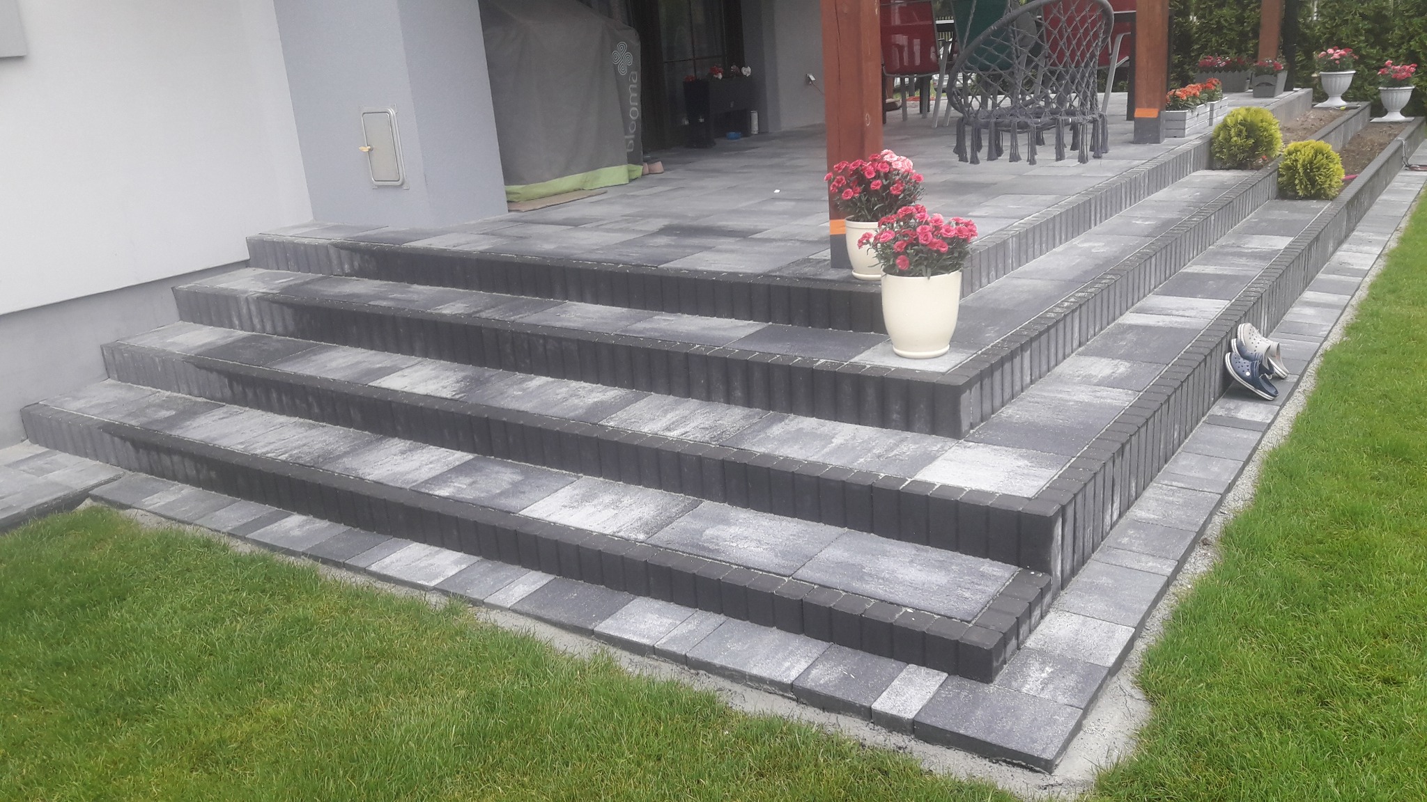 Schody wejściowe do domu wyłożone szarą kostką brukową z ciemnymi obrzeżami, donice z różowymi kwiatami i para klapek na trawniku.