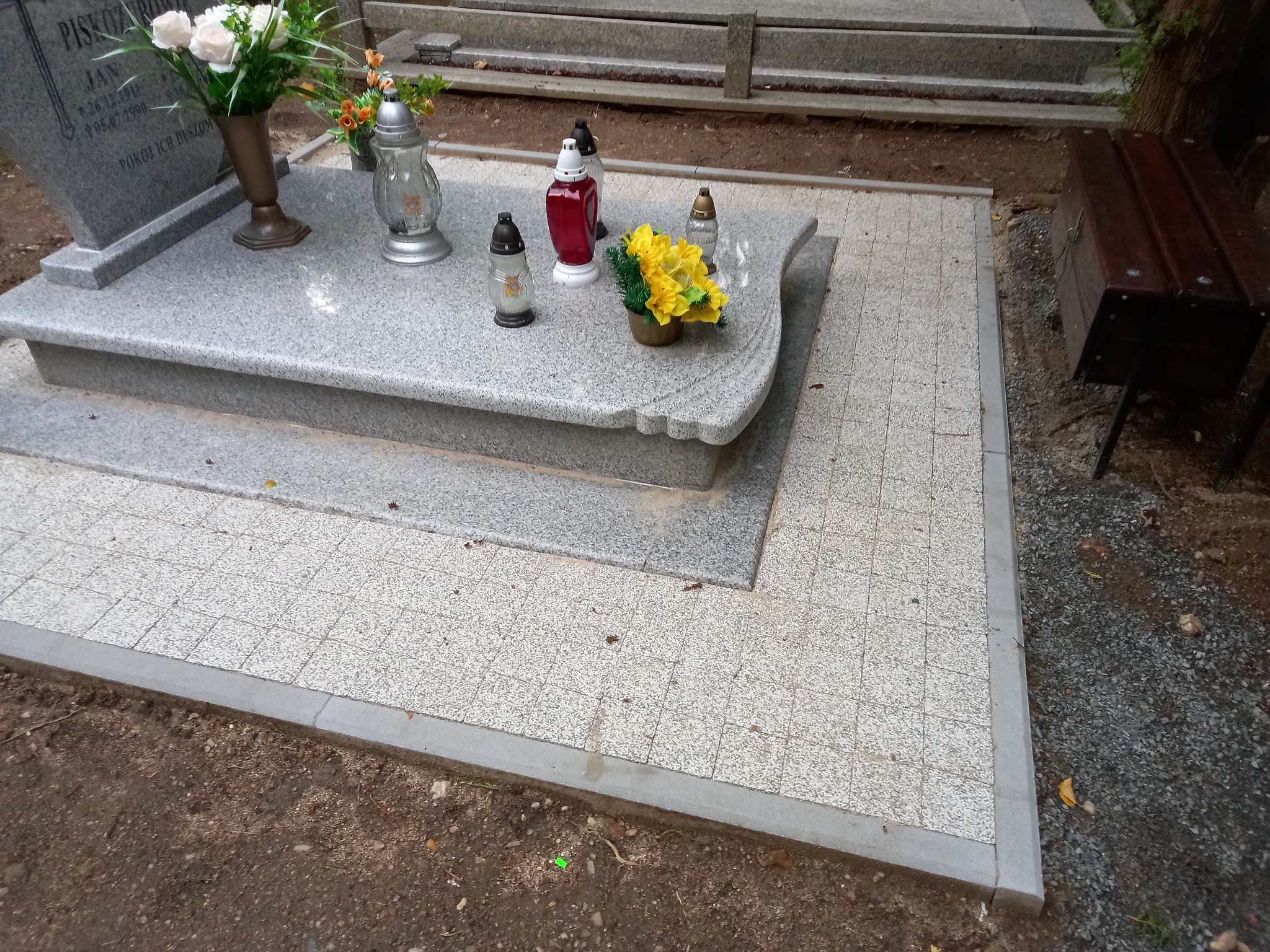 Wyłożenie kostką wokół nagrobka z kwiatami i zniczami, widoczna drewniana ławka.