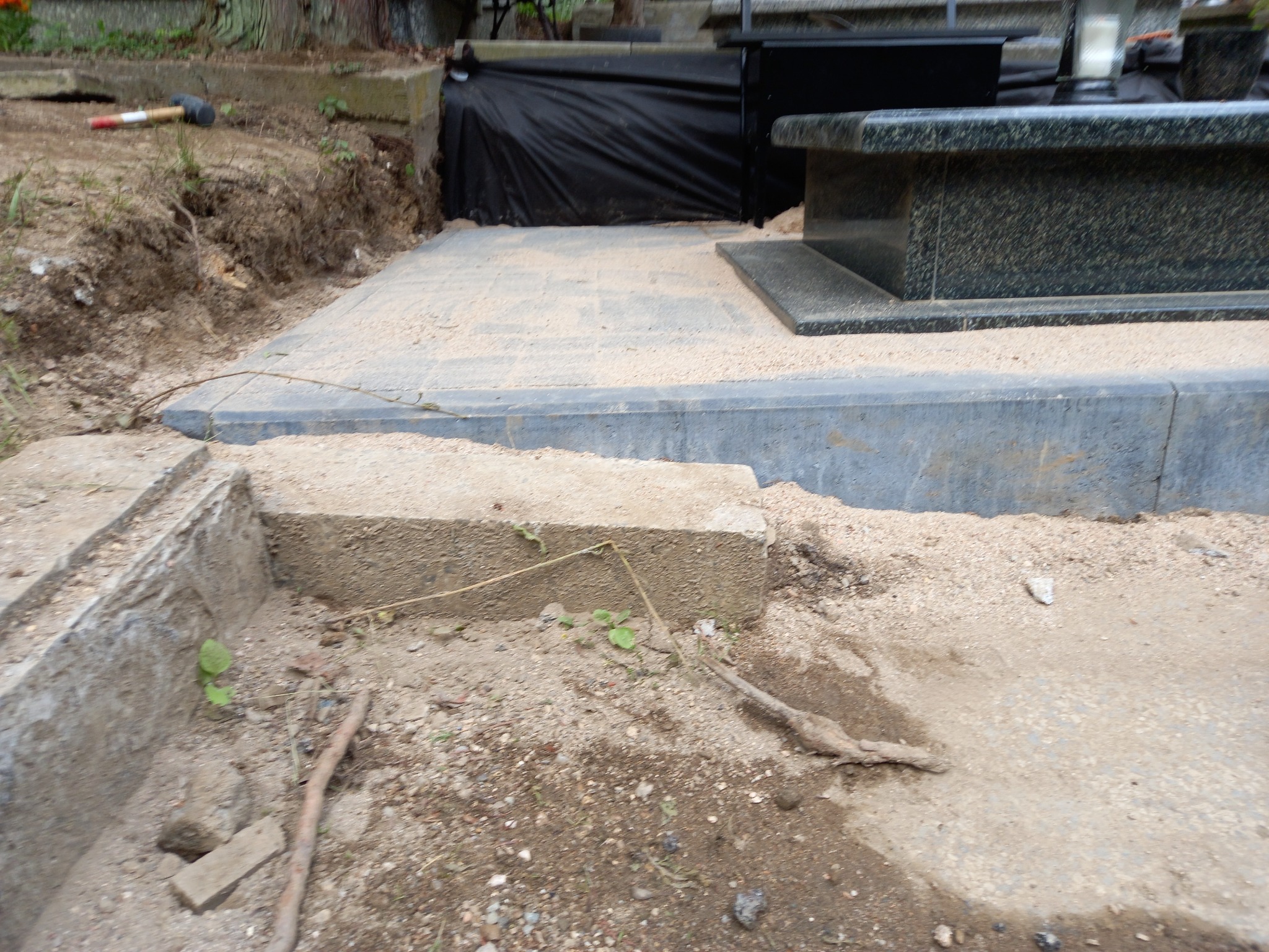 Przygotowanie terenu pod nagrobek w Jeleniej Górze: wyrównana powierzchnia z piaskiem, widoczny fragment nagrobka z ciemnego kamienia, obok leży gumowy młotek.