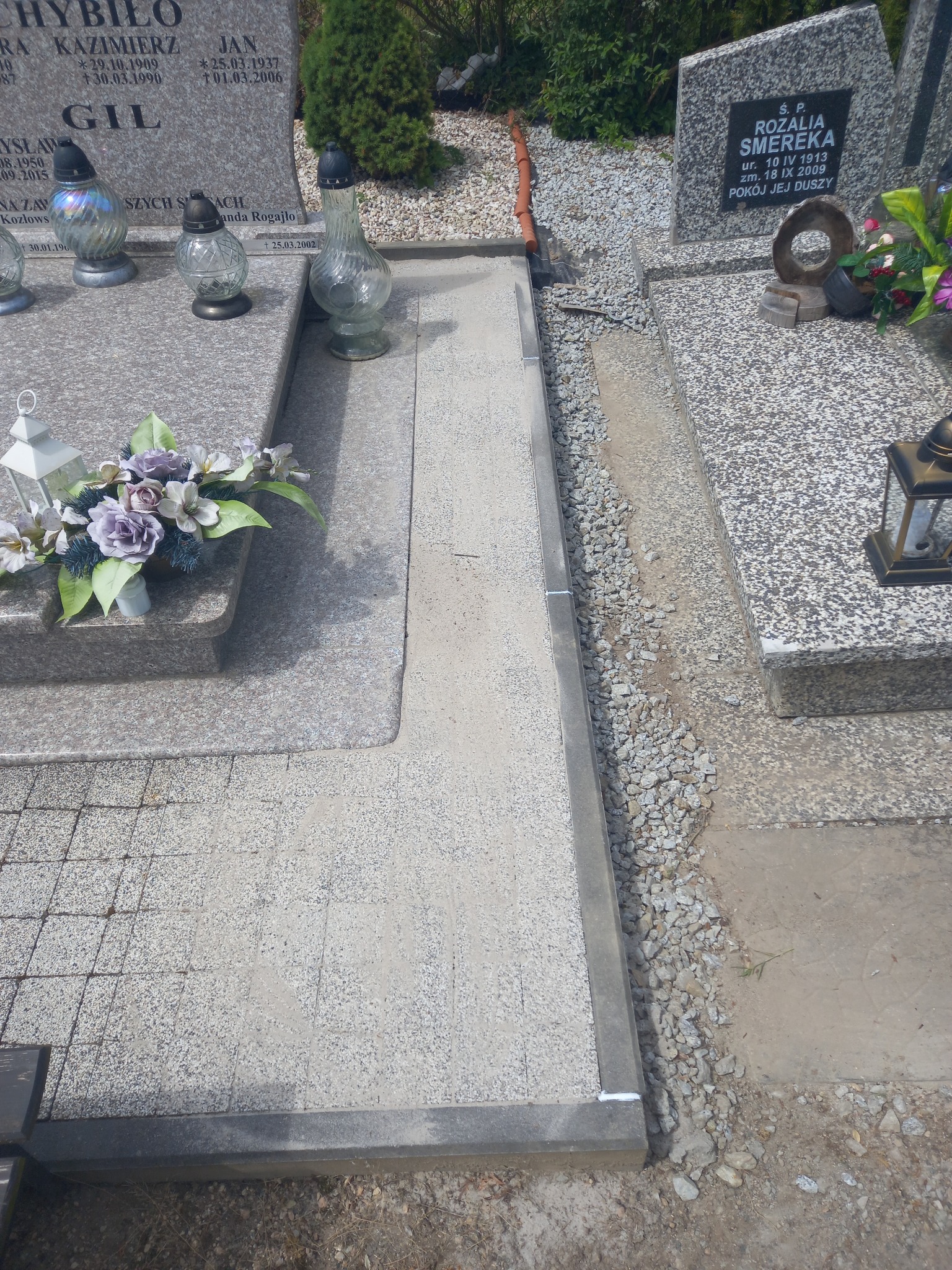 Wyłożona jasnoszarą kostką ścieżka między nagrobkami na cmentarzu, obrzeża wykończone metalowymi profilami, widoczne kwiaty i znicze na nagrobkach.