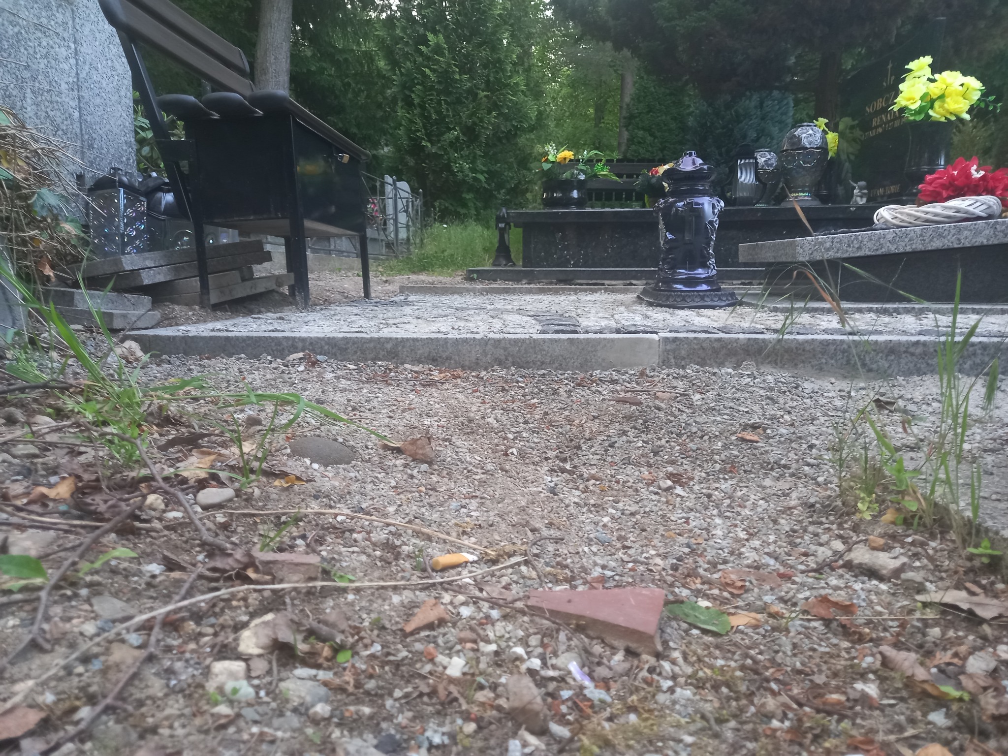 Nagrobek z kwiatami i ławką na cmentarzu, z ziemią, liśćmi i niedopałkiem papierosa na pierwszym planie.