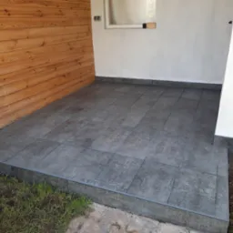 Wykończony taras z szarych, kwadratowych płytek ceramicznych, z drewnianą ścianą po lewej i białą po prawej stronie, z widocznym fragmentem trawnika u dołu.