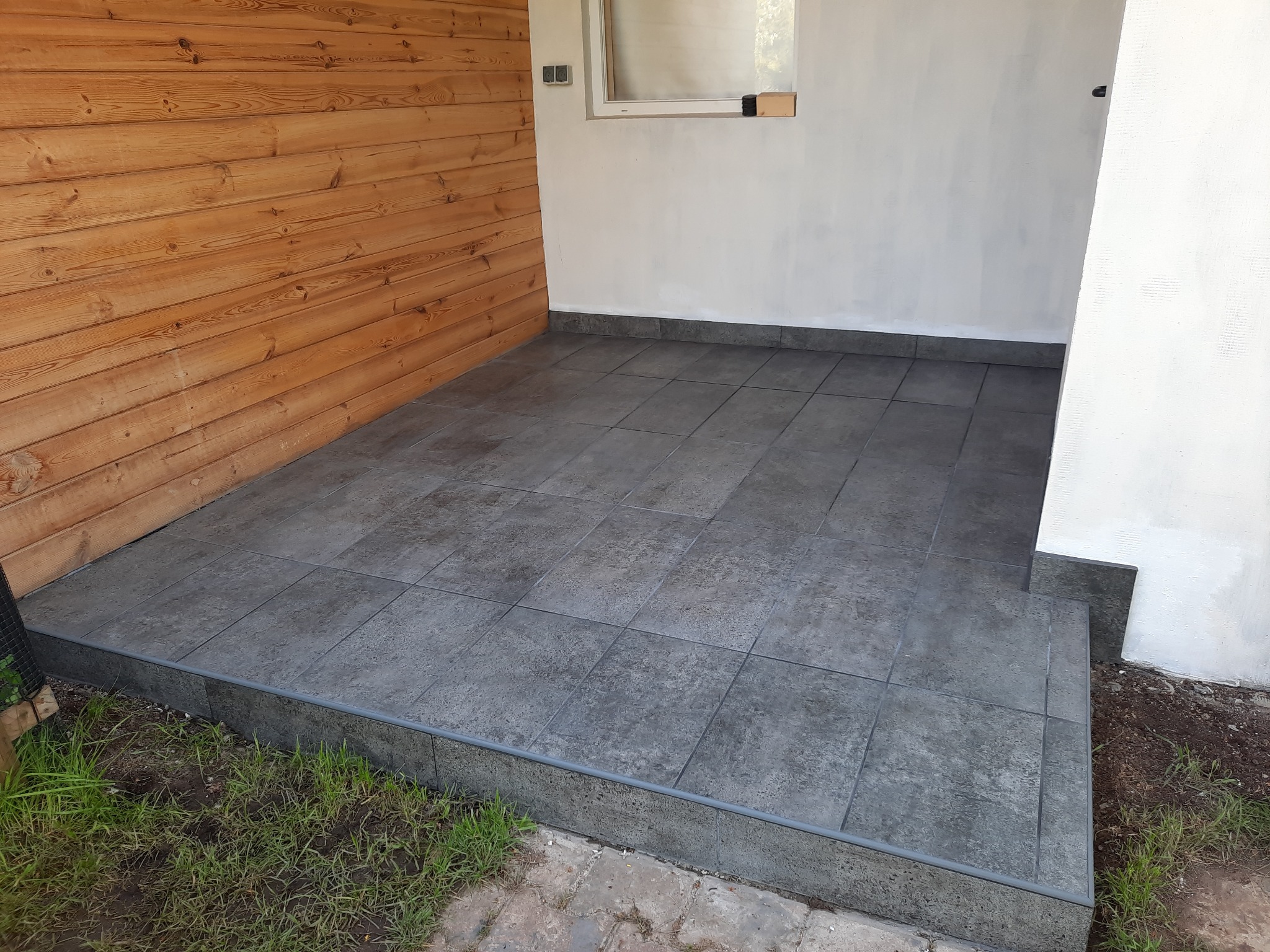 Wykończony taras z szarych, kwadratowych płytek ceramicznych, z drewnianą ścianą po lewej i białą po prawej stronie, z widocznym fragmentem trawnika u dołu.
