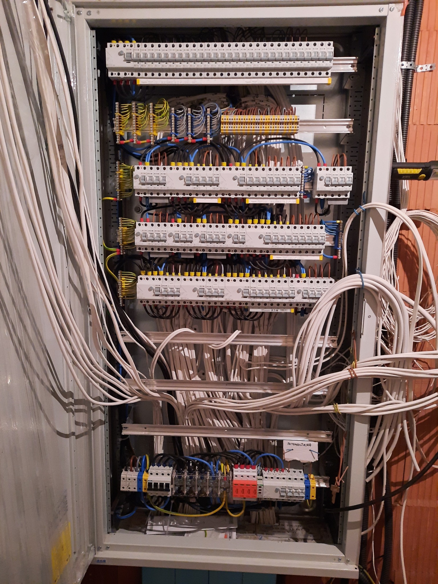 Otwarta skrzynka elektryczna z licznymi przewodami, bezpiecznikami i złączkami, widoczne kable w różnych kolorach, ułożone wewnątrz metalowej obudowy. Na dole widoczna kartka z napisem 'FOTONOLTAIKA'.