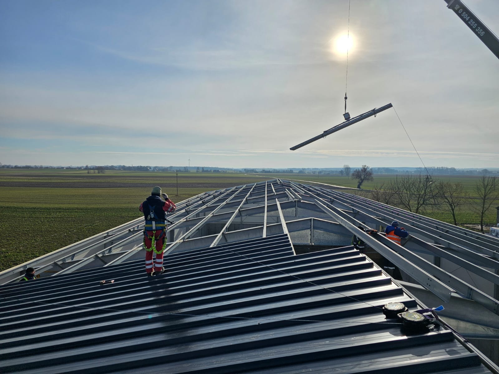 Montaż stalowej konstrukcji dachu hali z użyciem dźwigu, pracownicy w strojach roboczych na tle pól uprawnych i błękitnego nieba z promieniami słońca.