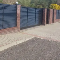 Nowoczesne ogrodzenie panelowe z poziomymi lamelami w kolorze antracytowym, osadzone na słupkach z czerwonej cegły, widoczne z zewnątrz posesji na tle zieleni drzew.