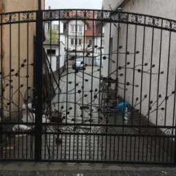 Czarna, metalowa brama wjazdowa z dekoracyjnym motywem gałęzi z liśćmi, widziana z zewnątrz posesji, z budynkami w tle.