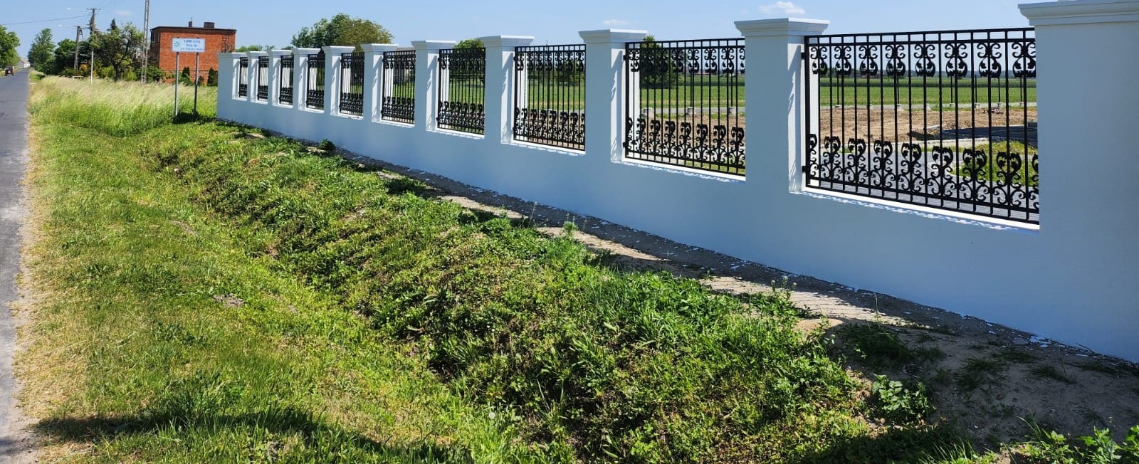 Nowoczesne ogrodzenie z białymi murowanymi słupkami i czarnymi metalowymi przęsłami z ozdobnymi elementami, widok z poziomu trawnika.