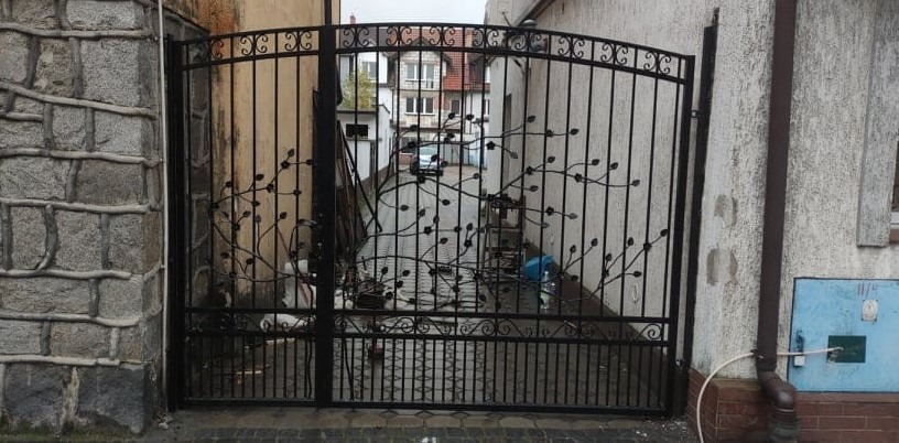 Czarna, metalowa brama wjazdowa z dekoracyjnym motywem gałęzi z liśćmi, widziana z zewnątrz posesji, z budynkami w tle.