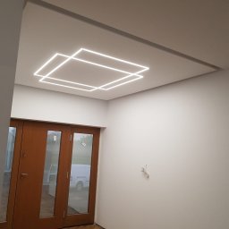 GOLD MAJSTER sp. z o.o. - Minimalistyczne wnętrze z parkietem w jodełkę i podwieszonym sufitem z geometrycznym oświetleniem LED w kształcie kwadratów, widoczne drzwi dwuskrzydłowe z przeszkleniami i biała ściana...