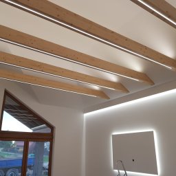 GOLD MAJSTER sp. z o.o. - Nowoczesne wnętrze z drewnianymi belkami stropowymi z wbudowanym oświetleniem LED i podświetleniem LED na ścianach, widoczne okno o nietypowym kształcie i drabina malarska.