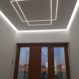 GOLD MAJSTER sp. z o.o. - Biały sufit z geometrycznym wzorem z ledowych linii, widoczne drewniane drzwi z szybami odbijającymi krajobraz.