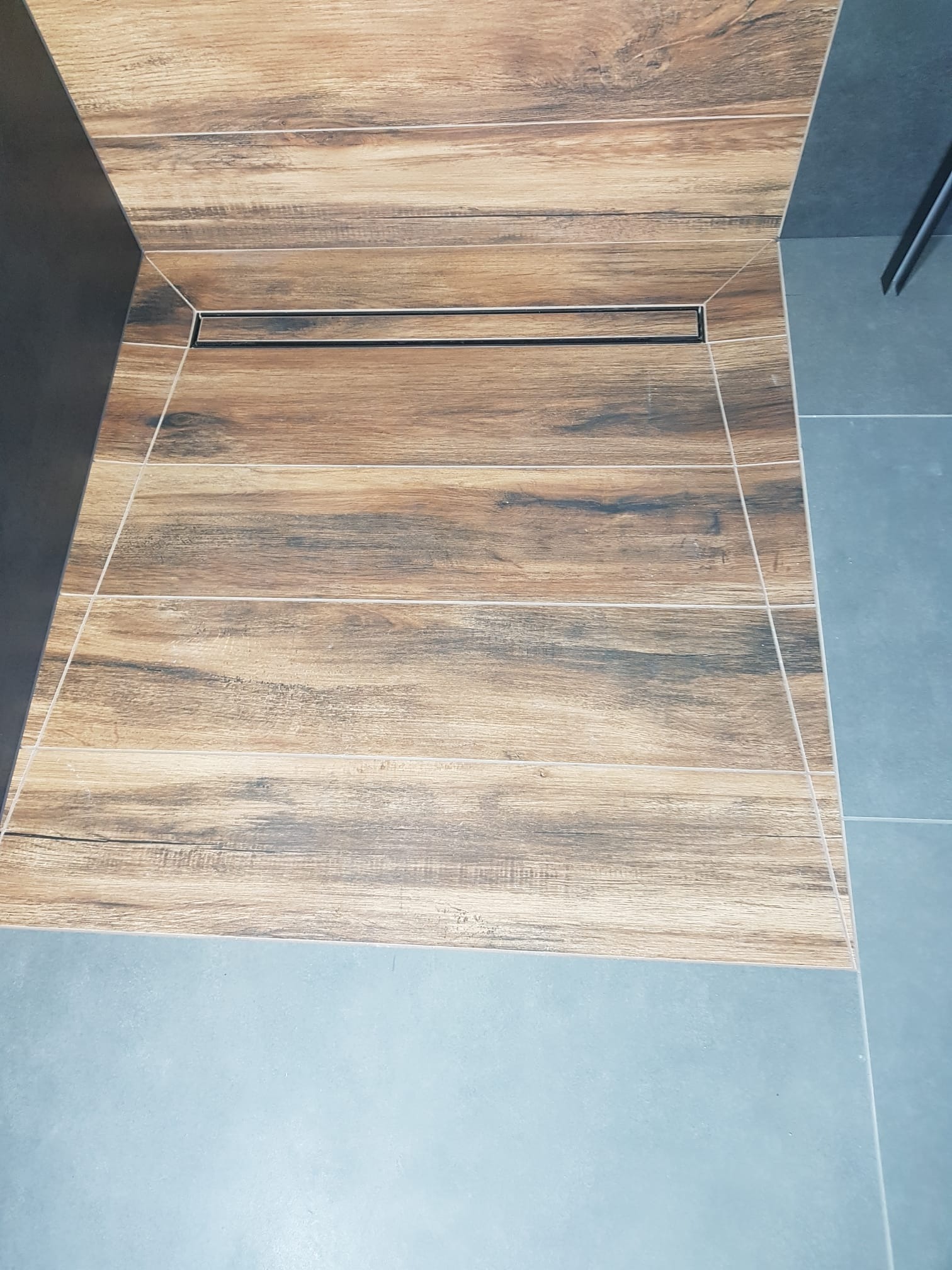 Płytki imitujące drewno na podłodze kabiny prysznicowej z odpływem liniowym, połączone z szarymi płytkami podłogowymi.