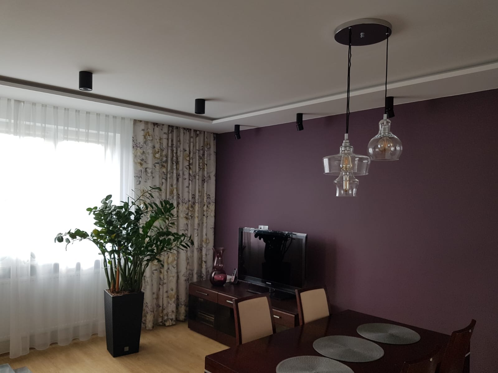 Nowoczesne wnętrze pokoju z fioletową ścianą, oświetleniem punktowym na suficie i wiszącymi lampami nad stołem. W rogu pokoju stoi duża roślina w czarnej donicy.