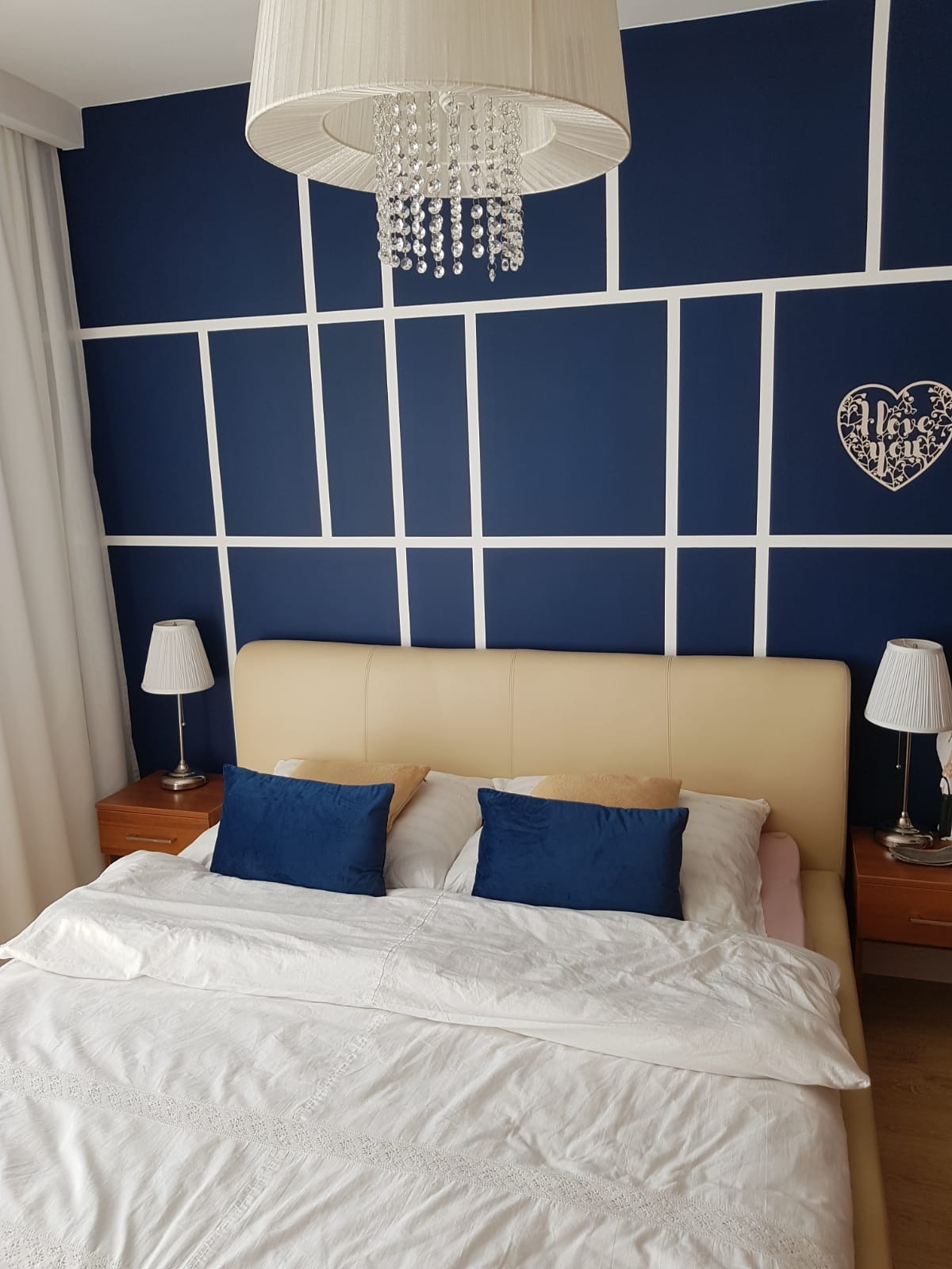 Elegancka sypialnia z granatową ścianą zdobioną białymi listwami, kryształowym żyrandolem i drewnianą dekoracją 'I love you', beżowe łóżko z niebieskimi poduszkami, dwie lampki nocne.