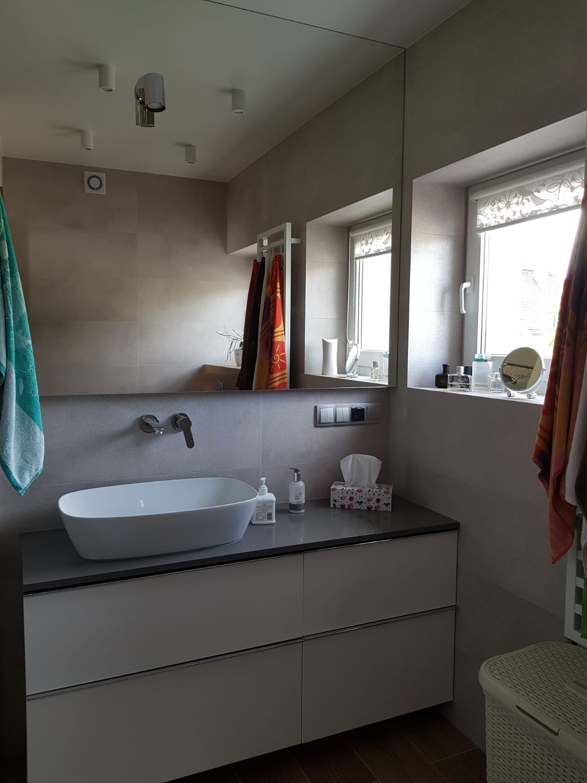 Nowoczesna łazienka z owalną umywalką nablatową, szarą szafką pod umywalką, dużym lustrem, oświetleniem punktowym i akcesoriami łazienkowymi na parapecie okna.
