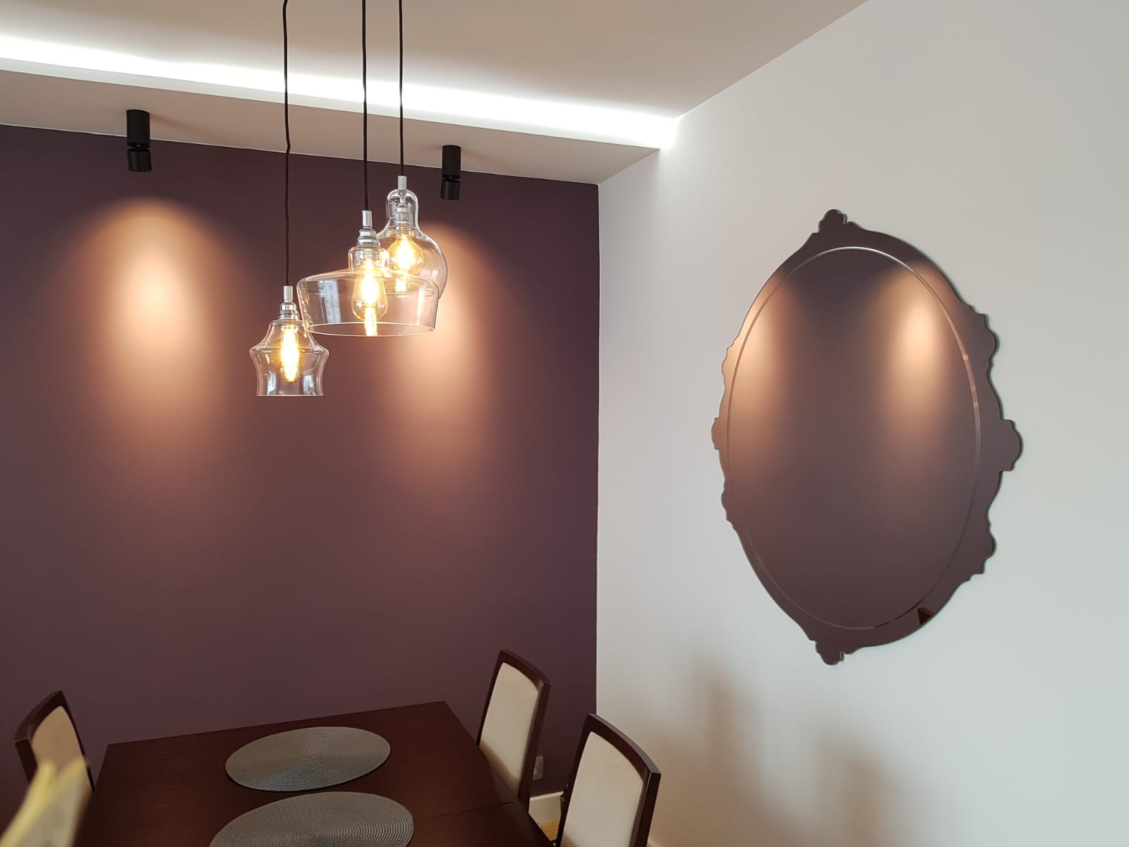 Jadalnia z ciemnofioletową ścianą, nowoczesną lampą wiszącą z trzema szklanymi kloszami, owalnym lustrem w ozdobnej ramie na białej ścianie oraz drewnianym stołem z dwoma okrągłymi podkładkami.