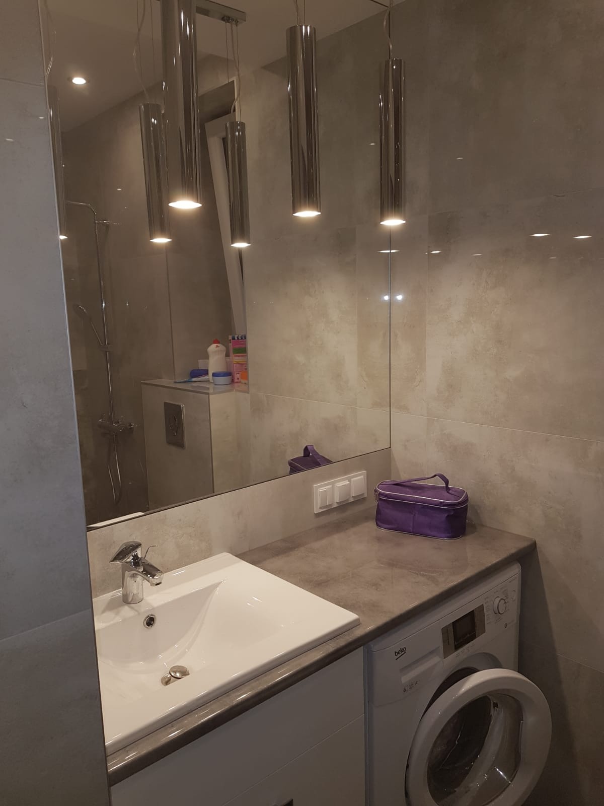 Nowoczesna łazienka z beżowymi płytkami, podwieszaną umywalką, pralką pod blatem i wiszącymi lampami tubowymi odbijającymi się w dużym lustrze.