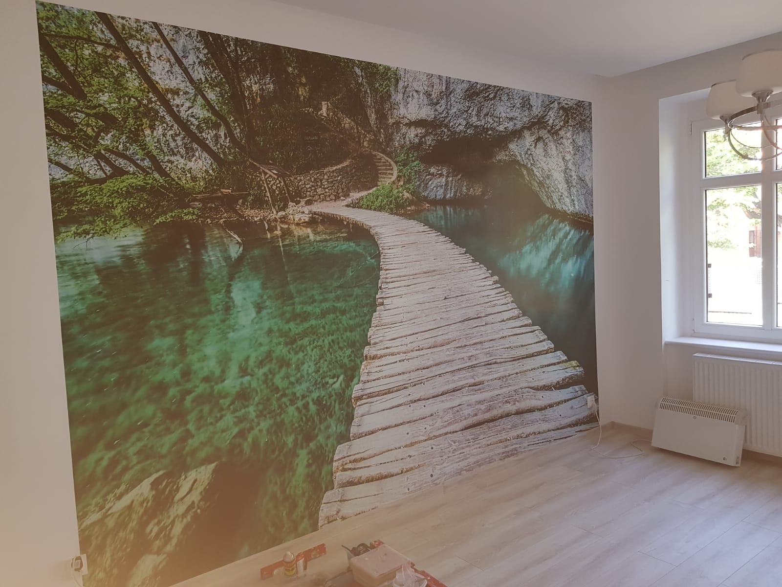 Fototapeta z motywem drewnianego mostu nad krystalicznie czystą wodą w lesie, naklejona na ścianie w jasnym pokoju z oknem i grzejnikiem.