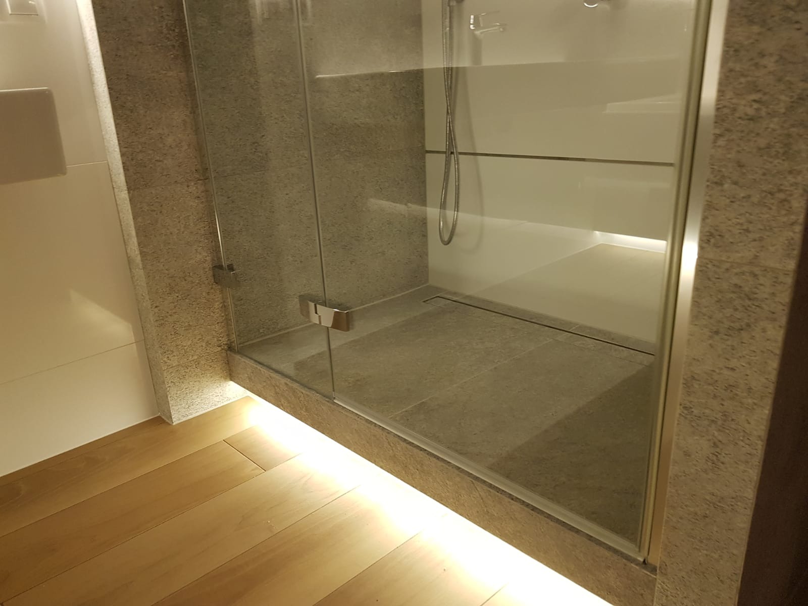 Nowoczesna kabina prysznicowa typu walk-in z odpływem liniowym, wykończona płytkami w odcieniach szarości i podświetleniem LED u podstawy, widoczna drewniana podłoga.