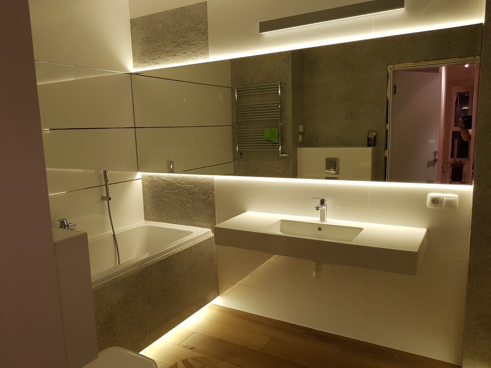 Nowoczesna łazienka z podświetleniem LED: wanna obudowana płytkami imitującymi kamień, biała umywalka nablatowa, lustro z oświetleniem, drewniana podłoga.