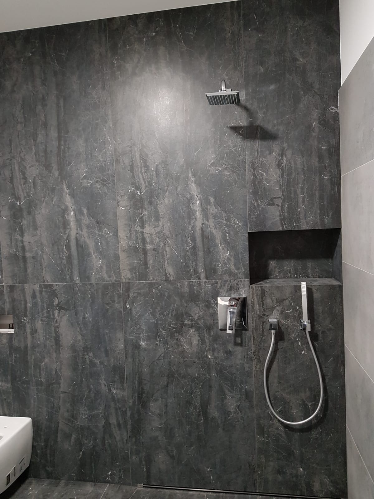 Wykończona łazienka z ciemnymi, marmurkowymi płytkami, deszczownicą, baterią podtynkową i wnęką z prysznicem ręcznym.