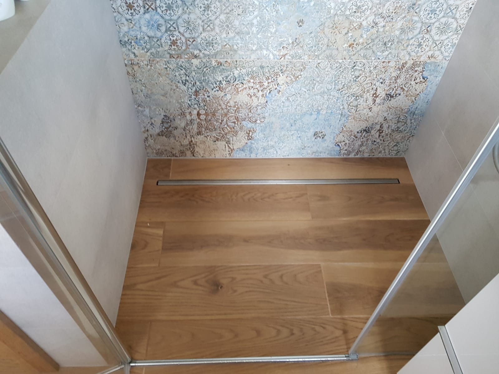 Kabina prysznicowa z odpływem liniowym w podłodze imitującej drewno i dekoracyjnymi płytkami na ścianie.