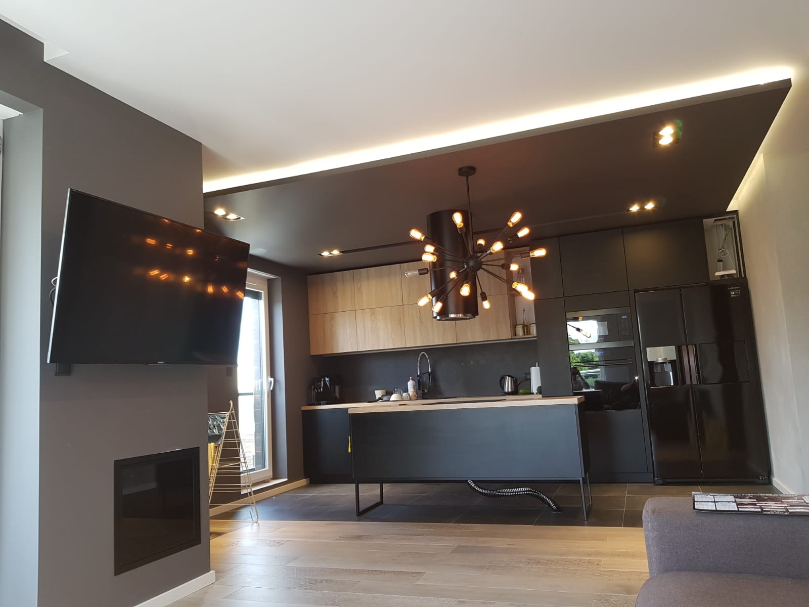 Nowoczesna kuchnia z czarnymi szafkami, drewnianymi akcentami i designerską lampą wiszącą z wieloma żarówkami, widok na salon z szarą ścianą i telewizorem.