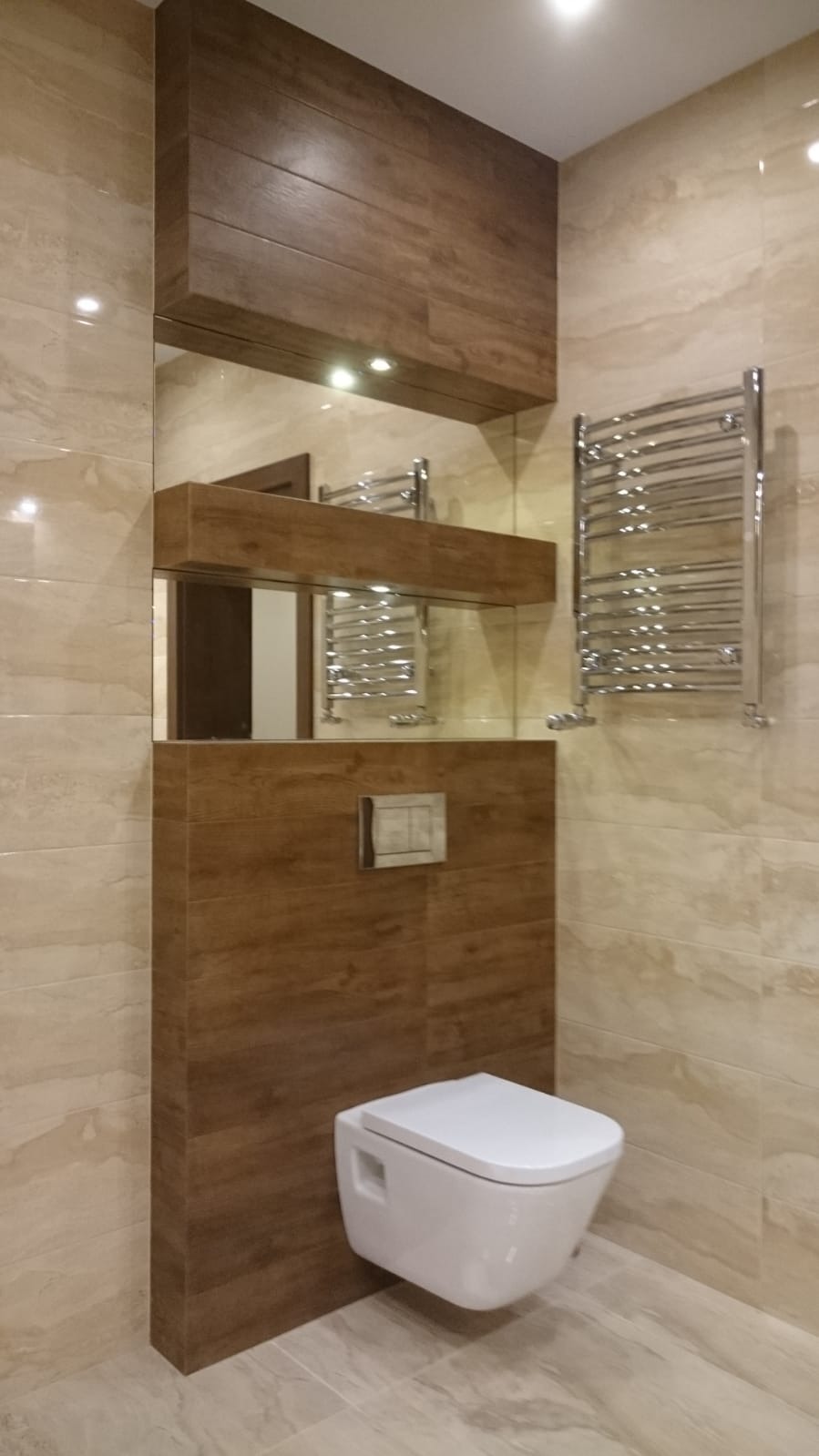 Nowoczesna łazienka z podwieszaną toaletą, drewnopodobnymi płytkami na zabudowie stelaża i szafkach, beżowe płytki imitujące marmur na ścianach, lustro z oświetleniem punktowym i grzejnik drabinkowy.