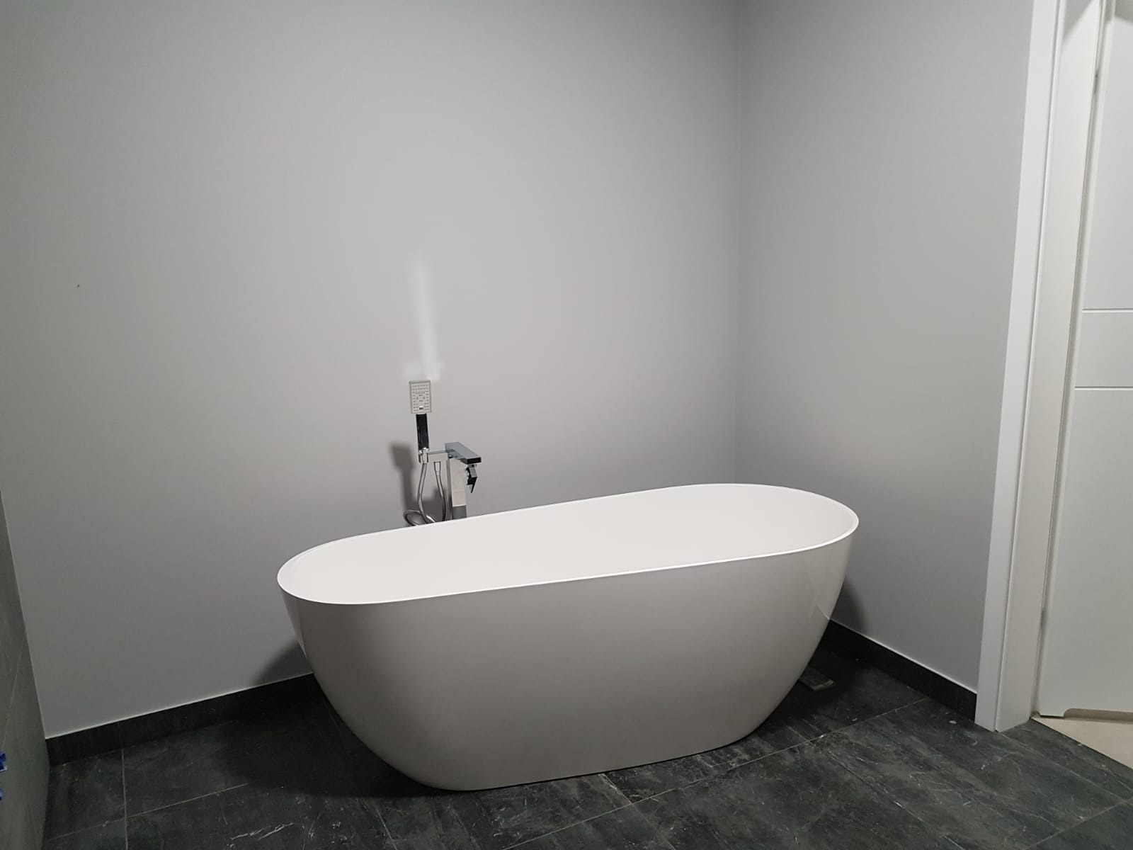 Nowoczesna, wolnostojąca, biała wanna owalna z chromowaną baterią i słuchawką prysznicową na tle szarej ściany i ciemnej, marmurkowej podłogi w łazience.