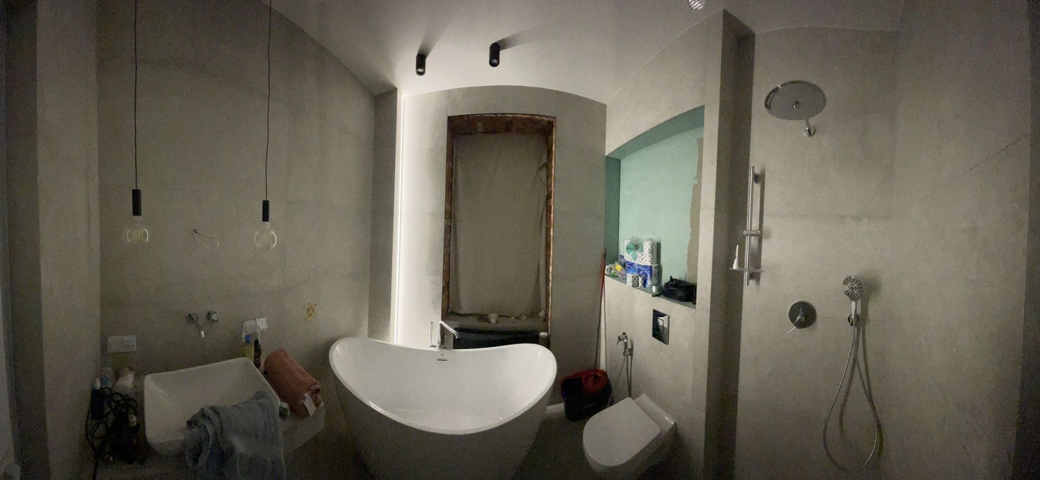 Łazienka w trakcie remontu: wolnostojąca wanna, surowa ściana z widocznymi niedoskonałościami, otwarty prysznic z deszczownicą i półka z akcesoriami łazienkowymi, wiszące żarówki na kablach.