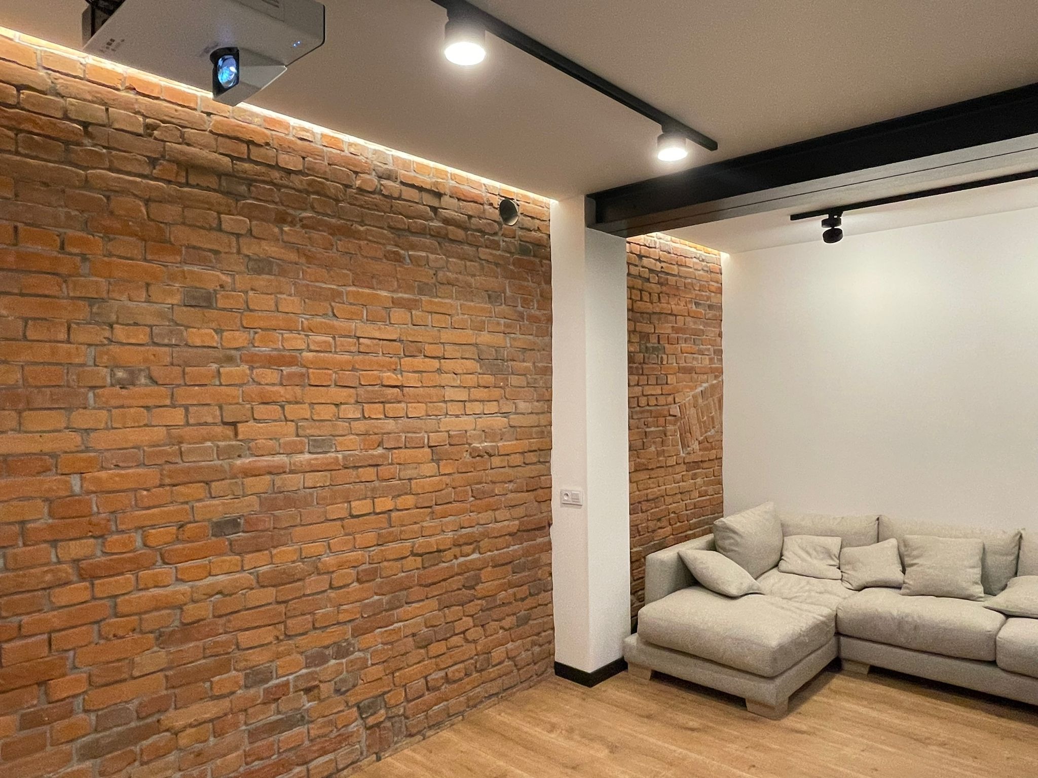 Salon z ceglaną ścianą, drewnianą podłogą i szarą sofą narożną, oświetlony reflektorami i listwą LED, z projektorem zamontowanym na suficie.