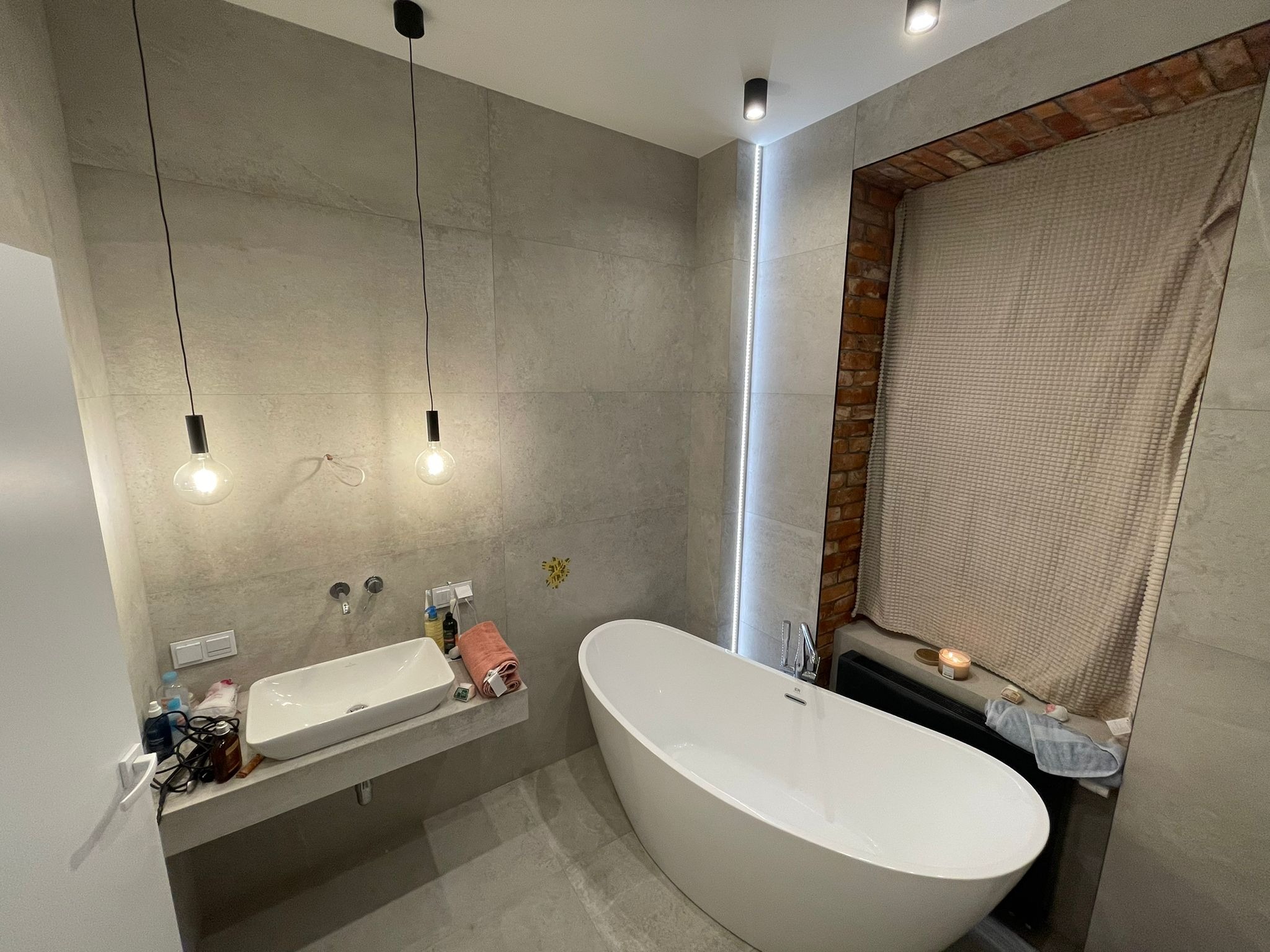 Nowoczesna łazienka z wanną wolnostojącą, umywalką nablatową na betonowym blacie, szarą glazurą i dekoracyjnym oświetleniem: wiszące żarówki i listwa LED przy ceglanym oknie.