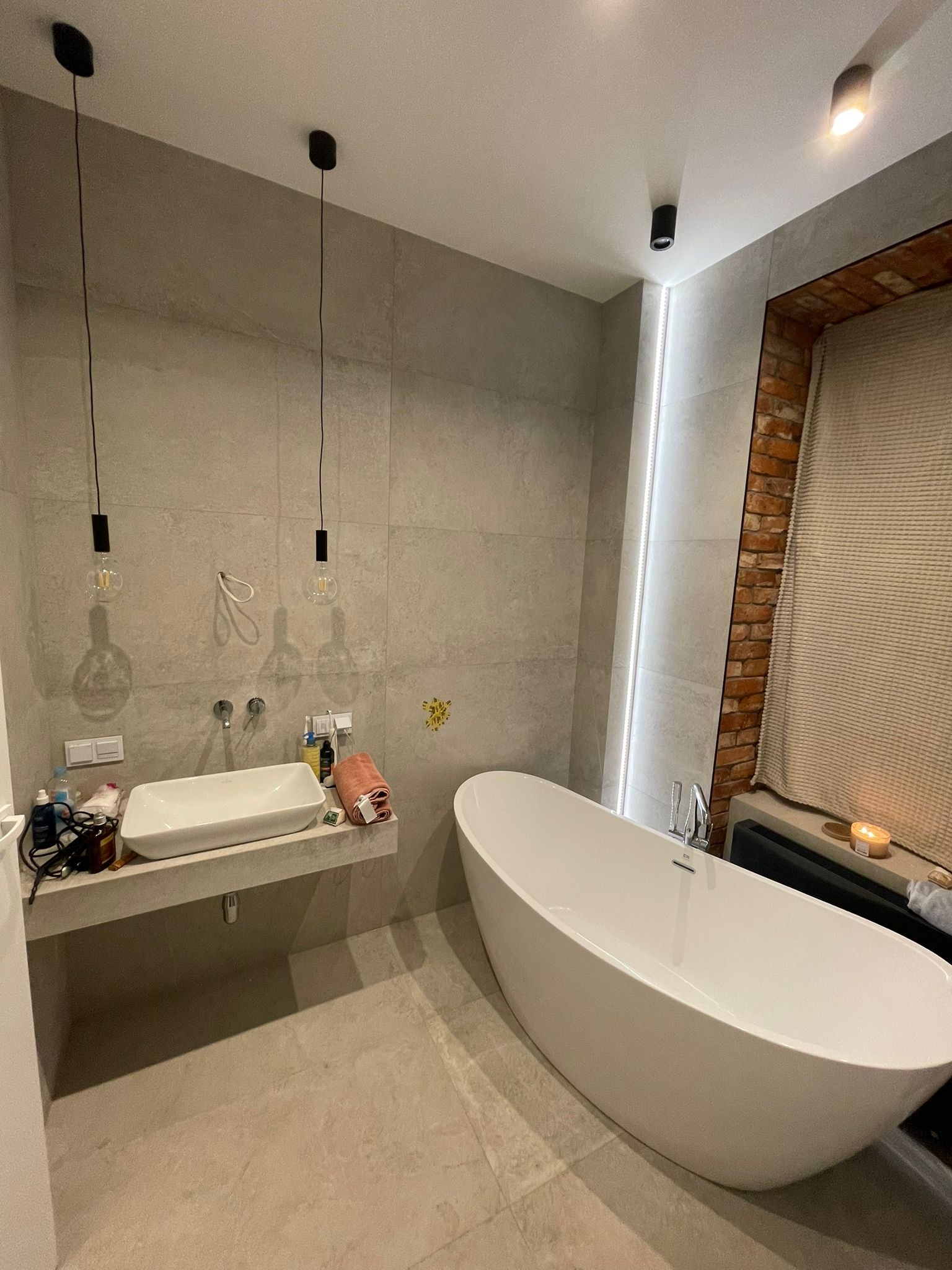 Nowoczesna łazienka z wolnostojącą białą wanną, szarą płytką imitującą beton, umywalką nablatową i dekoracyjnym oświetleniem, w tle fragment ściany z cegły.