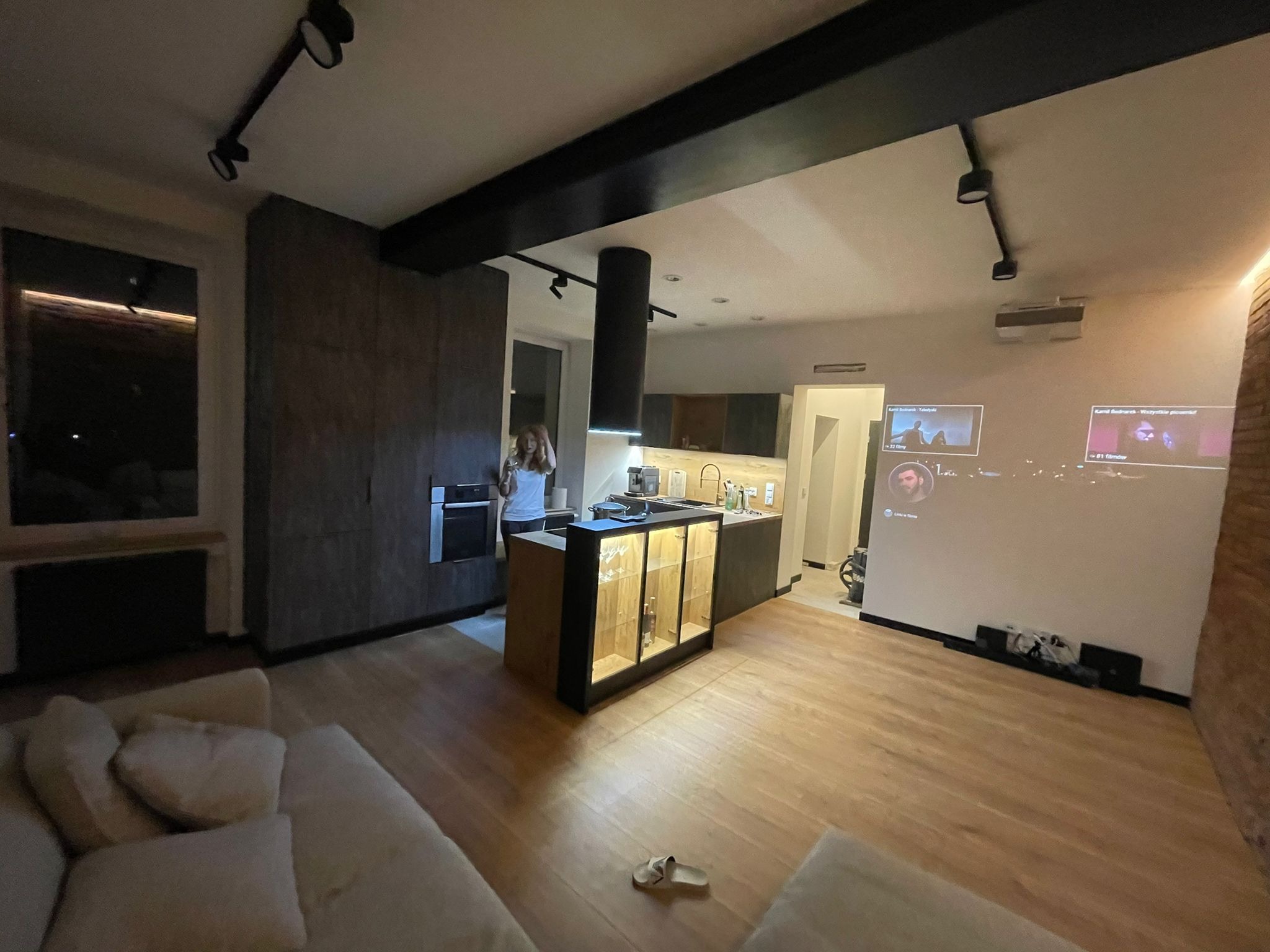 Nowoczesne wnętrze salonu z aneksem kuchennym, drewniana podłoga, szara ściana z fakturą betonu, czarna zabudowa kuchenna, wyspa z przeszkloną witryną, projektor wyświetlający obraz na ścianie.