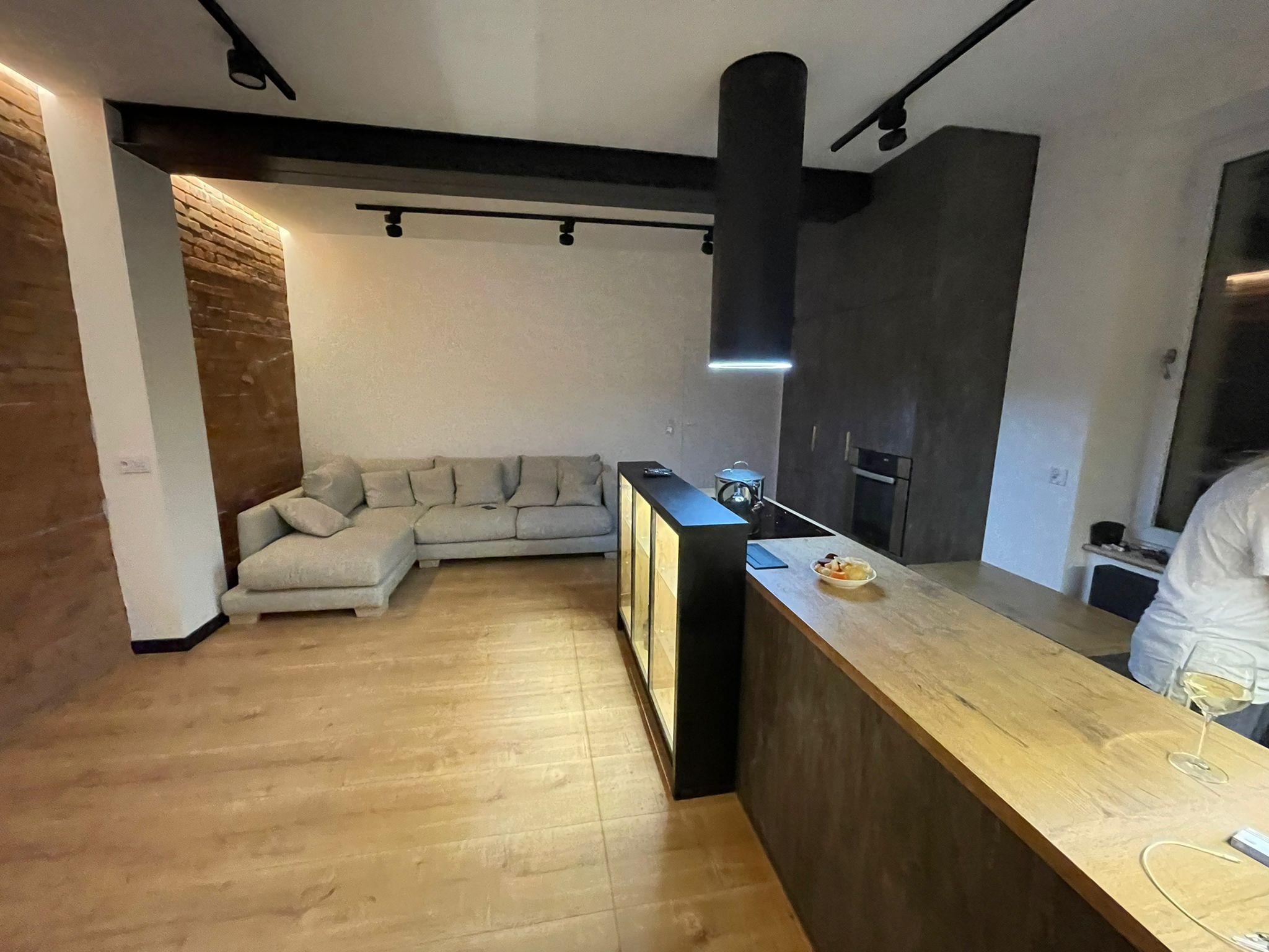 Nowoczesne wnętrze: salon z sofą narożną i aneksem kuchennym z drewnianym blatem, oświetlenie szynowe, ściana z cegły, czarny okap cylindryczny nad płytą grzewczą, podłoga z paneli imitujących drewno.