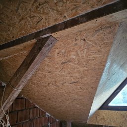 QMetters Sp. z o.o. - Poddasze z widocznymi drewnianymi belkami konstrukcyjnymi, płytami OSB, oknem trójkątnym i fragmentami murowanej ściany z czerwonej cegły, częściowo wypełnionej pianką izolacyjną.