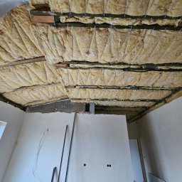 QMetters Sp. z o.o. - Poddasze w trakcie ocieplania wełną mineralną, widoczne drewniane belki i druty mocujące, fragment ściany z otworami pod instalację elektryczną, surowy stan wykończenia wnętrza