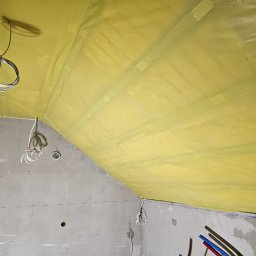 QMetters Sp. z o.o. - Żółta folia paroizolacyjna zamocowana do krokwi na poddaszu, widoczne kable elektryczne i fragment surowej ściany z naniesionymi oznaczeniami.