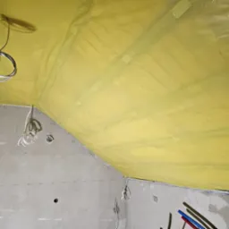 Żółta folia paroizolacyjna zamocowana do krokwi na poddaszu, widoczne kable elektryczne i fragment surowej ściany z naniesionymi oznaczeniami.