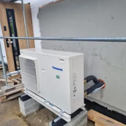 Zewnętrzna jednostka klimatyzacji Panasonic zamontowana na bloczkach betonowych przy ścianie budynku w trakcie prac wykończeniowych, widoczne rury połączeniowe z miedzianymi zaworami.