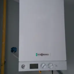 Wiszący na ścianie biały piec gazowy Viessmann Vitodens 100 z podłączonymi rurami gazową i wodną, widoczny manometr i pokrętła regulacyjne.