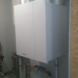 QMetters Sp. z o.o. - Biały piec grzewczy z widocznymi rurami instalacji, umieszczony w pomieszczeniu z izolacją z folii aluminiowej na suficie.