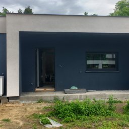 QMetters Sp. z o.o. - Nowoczesny dom jednorodzinny w trakcie wykończenia, z zamontowaną jednostką zewnętrzną pompy ciepła, widoczne niedokończone prace ziemne wokół budynku.