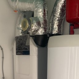 QMetters Sp. z o.o. - Wnętrze kotłowni z rekuperatorem Zehnder, widoczne rury wentylacyjne w izolacji aluminiowej, zbiornik akumulacyjny i instalacja hydrauliczna z zaworami.