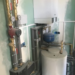 QMetters Sp. z o.o. - Kotłownia w trakcie montażu, widoczne rury miedziane z zaworami, manometry, pompa obiegowa Grundfos, zbiornik hydroforowy, bojler oraz instalacja w szarej izolacji na tle surowych, zagruntowanych...