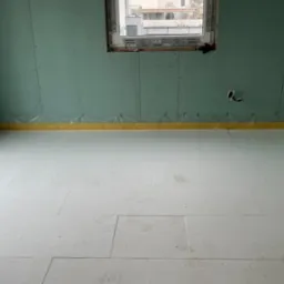 Poddasze w trakcie prac wykończeniowych: ściany obłożone płytami gipsowo-kartonowymi, zabezpieczone folią przy podłodze, na podłodze białe płyty izolacyjne z widocznymi śladami stóp, okno w tle.