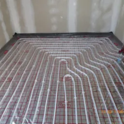 Układ rur grzewczych na podłodze z siatką montażową, przygotowany do zalania wylewką, widoczna butelka z wodą i fragment ściany w tle.