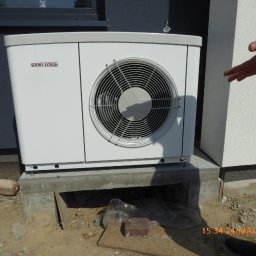 QMetters Sp. z o.o. - Biała pompa ciepła Stiebel Eltron na betonowym fundamencie, widoczna data wykonania instalacji, częściowo widoczna dłoń osoby wskazującej na urządzenie.