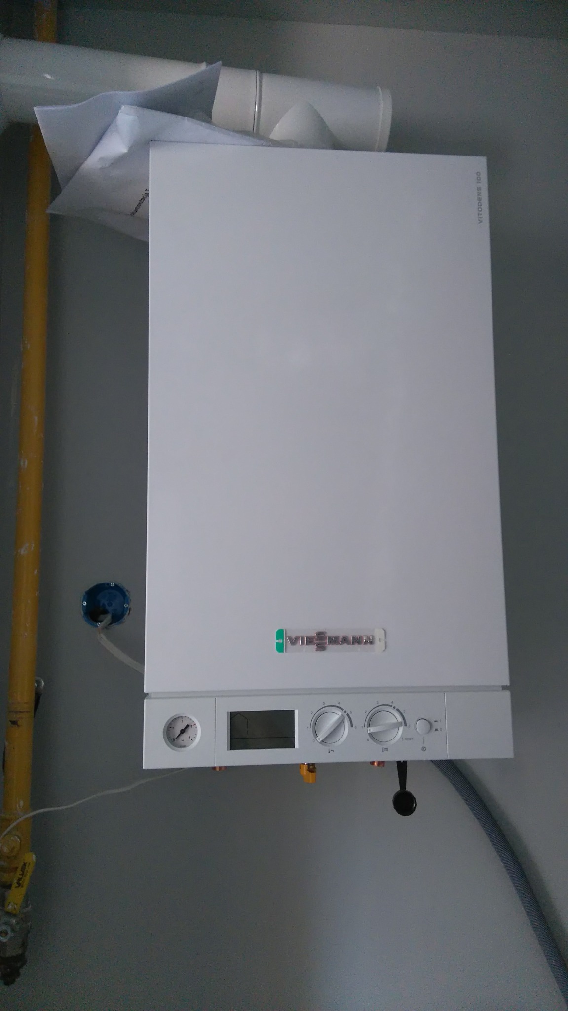 Wiszący na ścianie biały piec gazowy Viessmann Vitodens 100 z podłączonymi rurami gazową i wodną, widoczny manometr i pokrętła regulacyjne.
